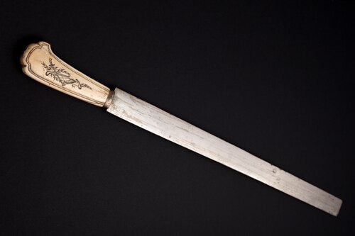 A long, straight chalaf (shechita knife) with an ornate, light-colored handle is displayed against a dark background. This particular knife dates back to the mid-18th century and is part of the Jewish Museum of Switzerland's collection.