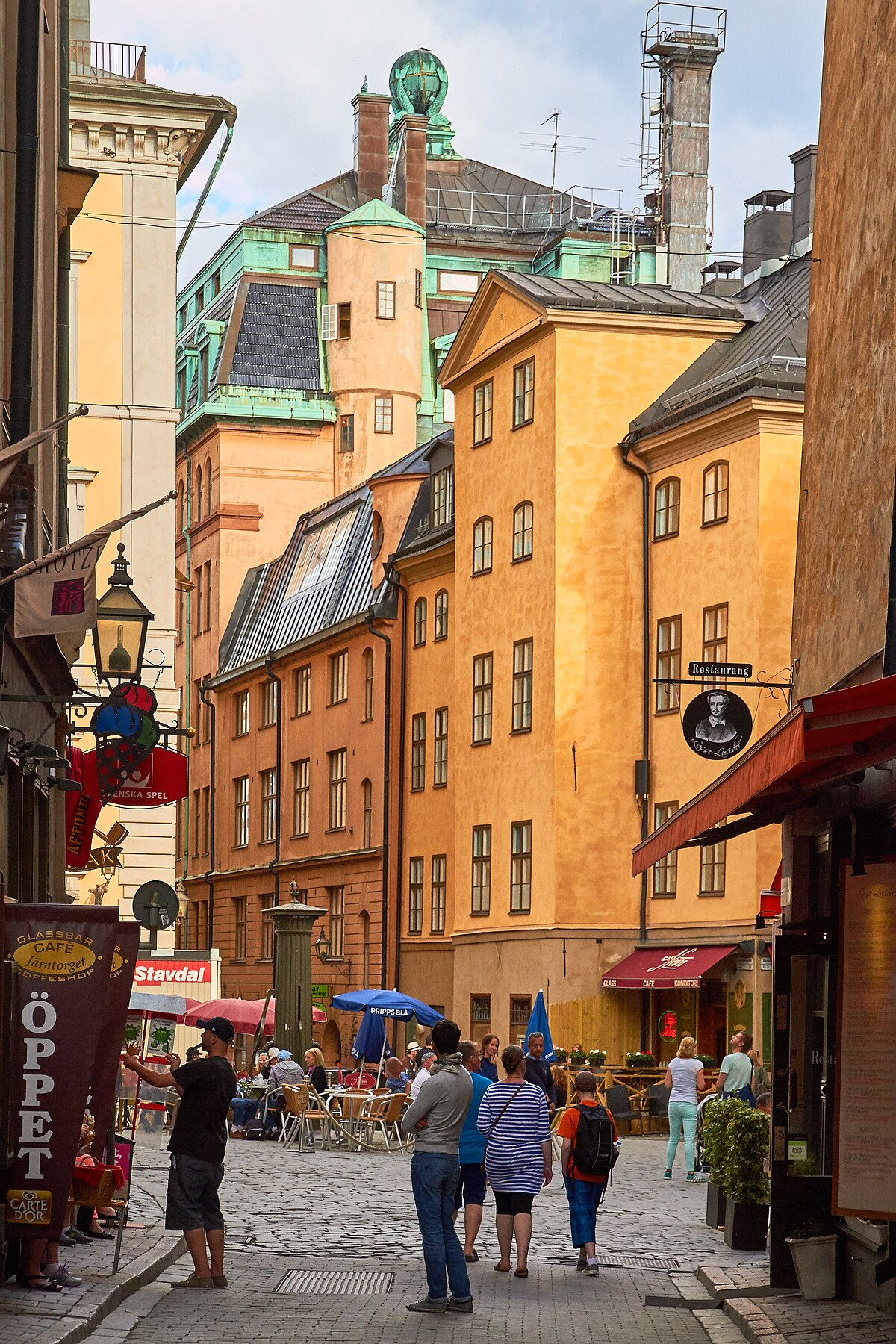 A street scene in Gamla Stan, Stockholm, features historic buildings with varied architecture and colors, including yellow, orange, and green roofs. People are walking and dining at outdoor cafes along the cobblestone street.