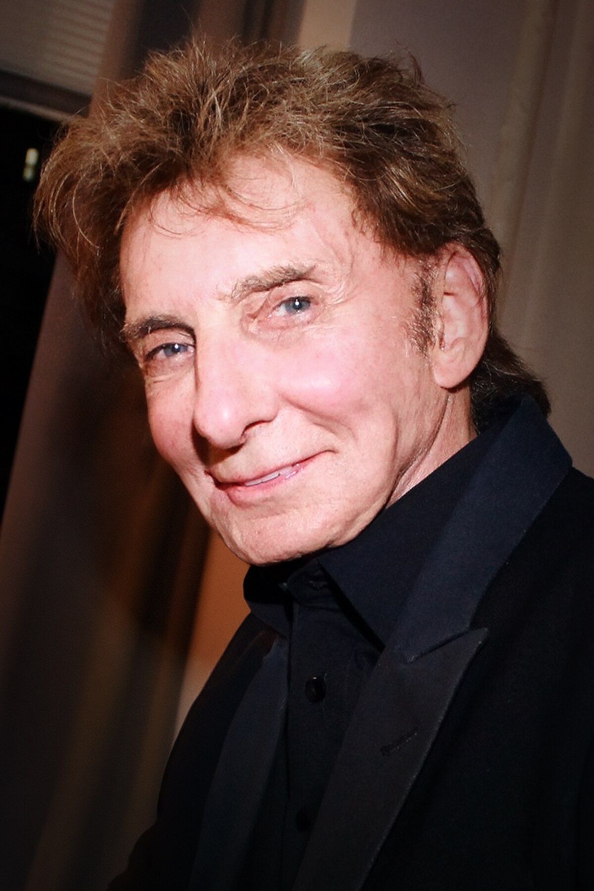 A portrait of Barry Manilow, smiling and looking towards the viewer. He has light brown hair and is wearing a black suit jacket and shirt.