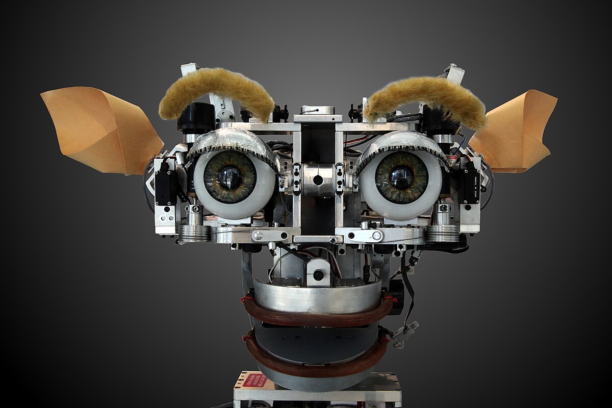 The robot head Kismet, developed in the 1990s, is shown with large, expressive eyes, mechanical components, and ear-like structures, designed to recognize and simulate emotions.
