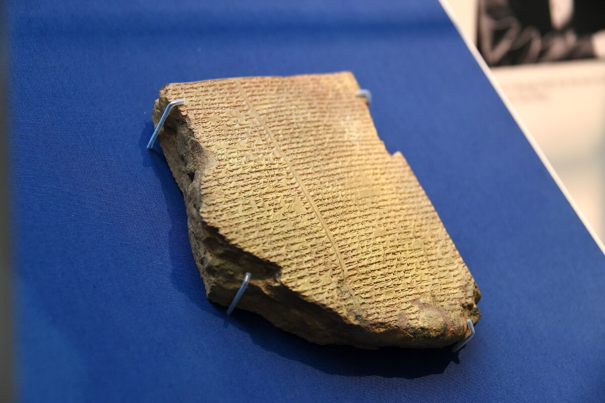 A fragment of Tablet XI, also known as the Flood Tablet, of the Epic of Gilgamesh, displayed in a museum. The cuneiform script is visible on the clay tablet, which is propped up on a blue surface.