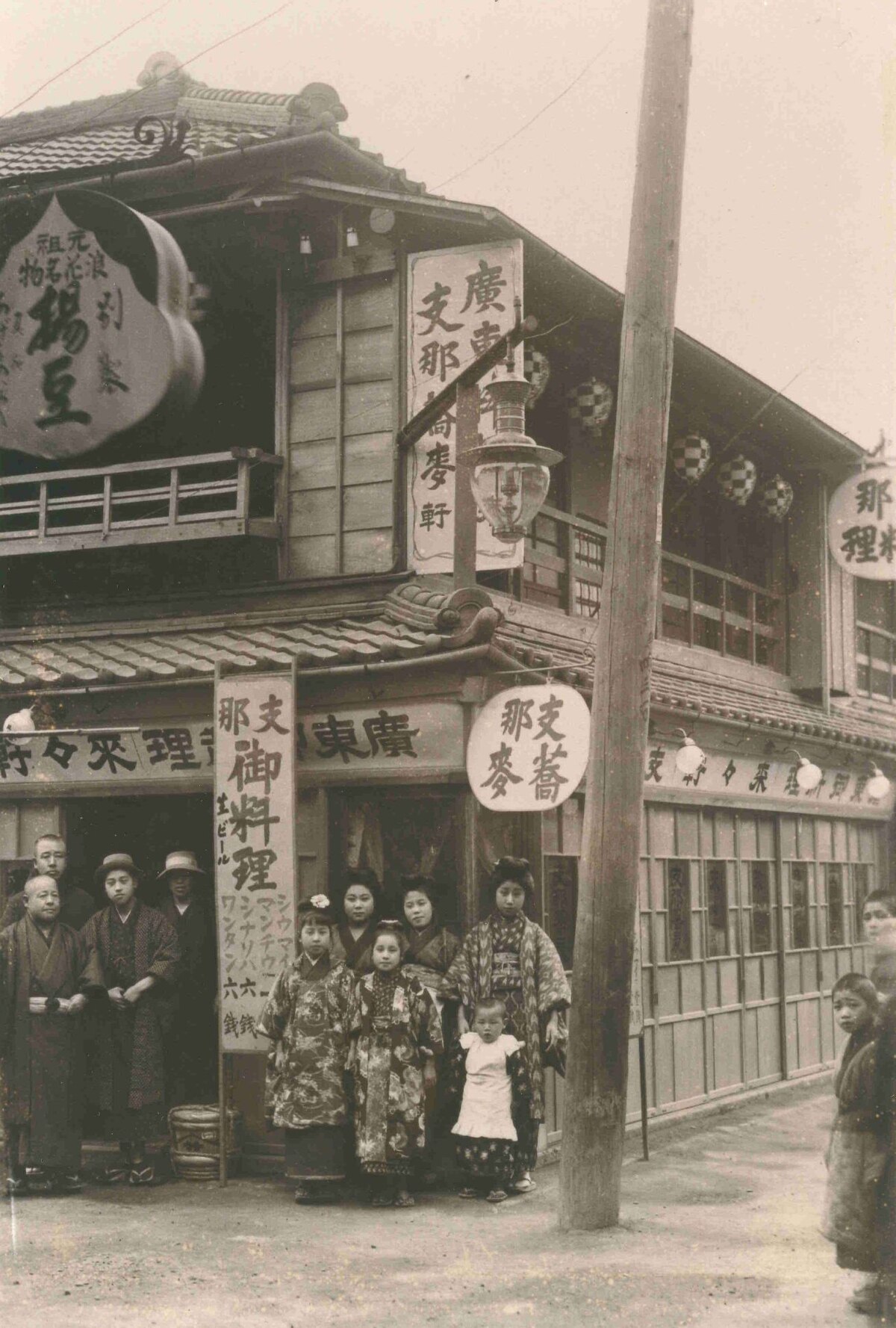 A vintage photograph of Rairaiken, the first ramen shop, founded in 1910 by Kan'ichi Ozaki in Asakusa, Tokyo. Several people, including women in traditional attire, stand outside the two-story building, which displays signs advertising "Chinese soba" (支那蕎麥) and "Cantonese cuisine" (廣東料理).