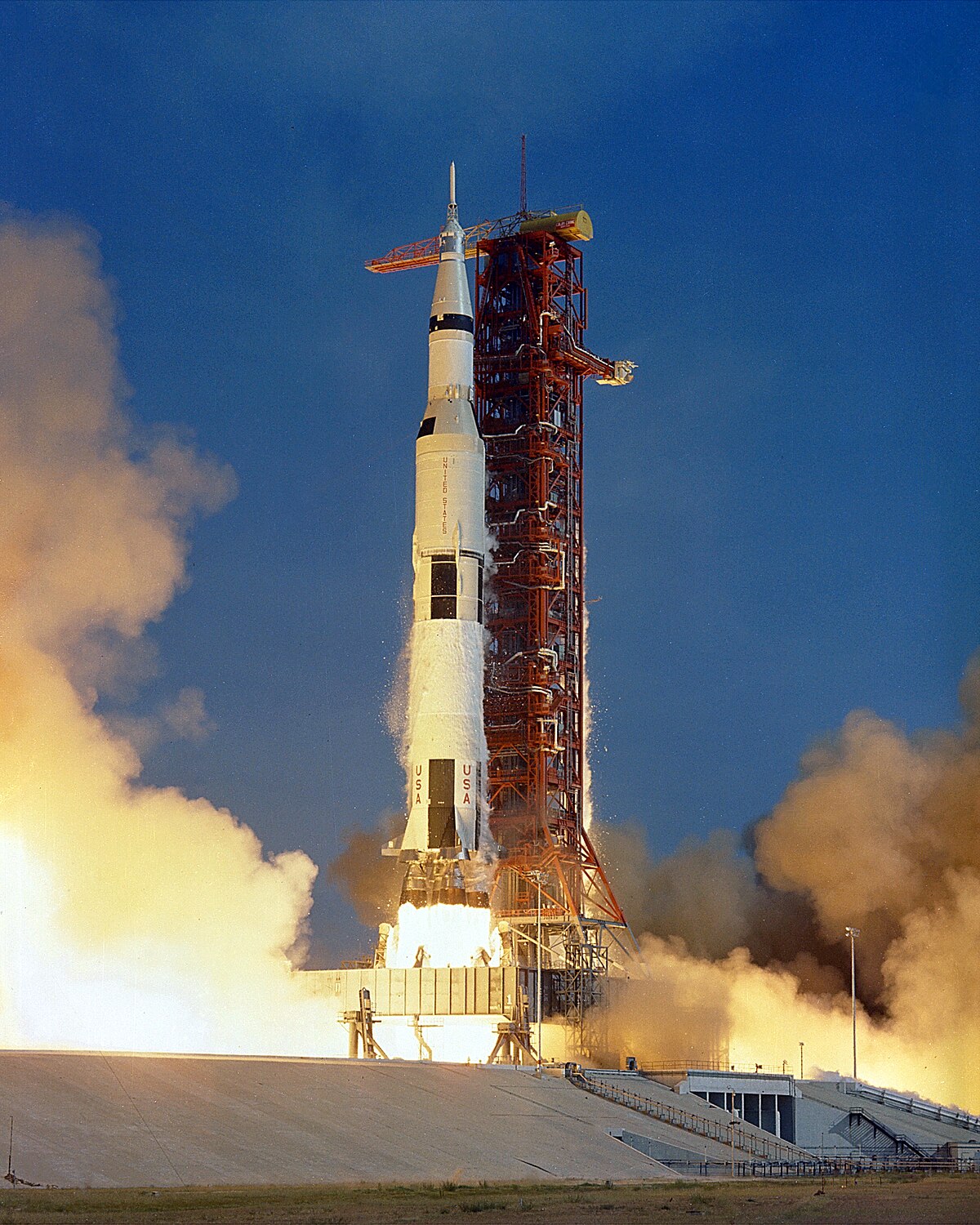 The Apollo 11 Saturn V rocket launching from Launch Complex 39A at Kennedy Space Center, surrounded by massive plumes of smoke and fire. The rocket is shown on its launch tower, with the "USA" flag visible on its side.