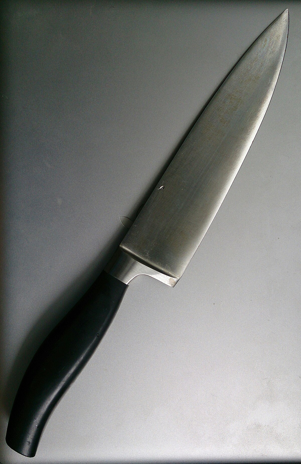 A chef's knife with a silver blade and a black handle, positioned diagonally on a gray surface. The blade shows signs of use with minor scratches and reflections.