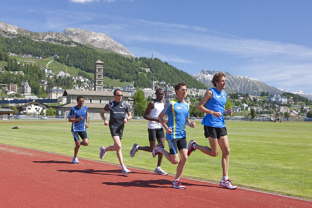 Athletes performing altitude training at the Swiss Olympic Training Base in St. Moritz, located at an elevation of 1,856 meters (6,089 feet). Five runners are on a red track with mountains and a town in the background.