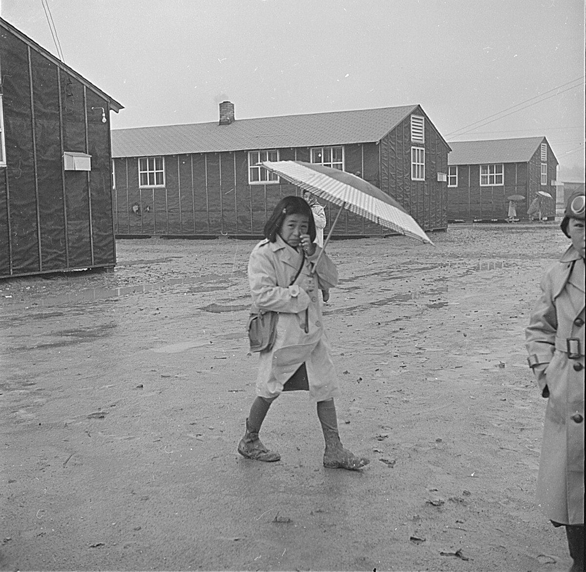 A young Japanese-American girl, wearing a coat and holding an umbrella, walks through a muddy internment camp in Arkansas in 1943. Wooden barracks are visible in the background.