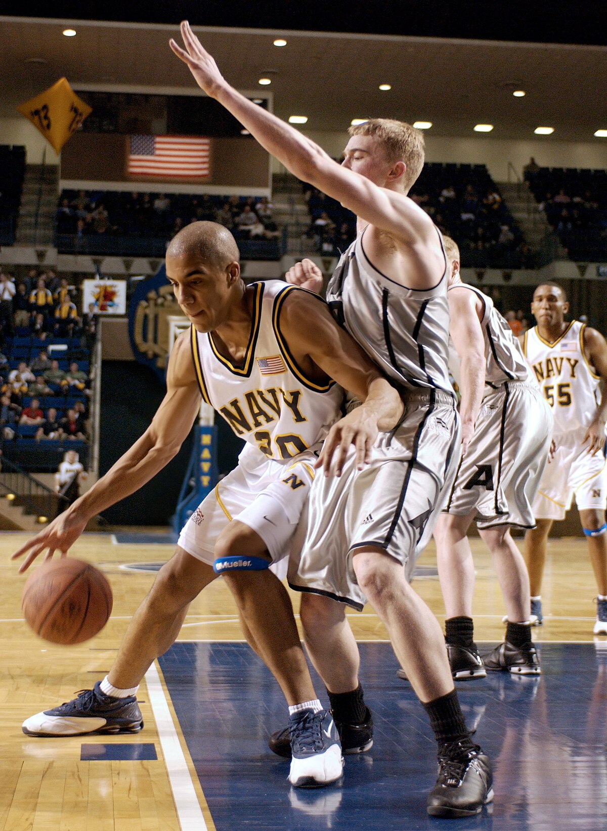 A Navy basketball player, wearing a white jersey with number 20, dribbles the ball while being defended by an Army player in a grey jersey during the 2004 Army vs. Navy Basketball game. Another Navy player with number 55 is visible in the background.