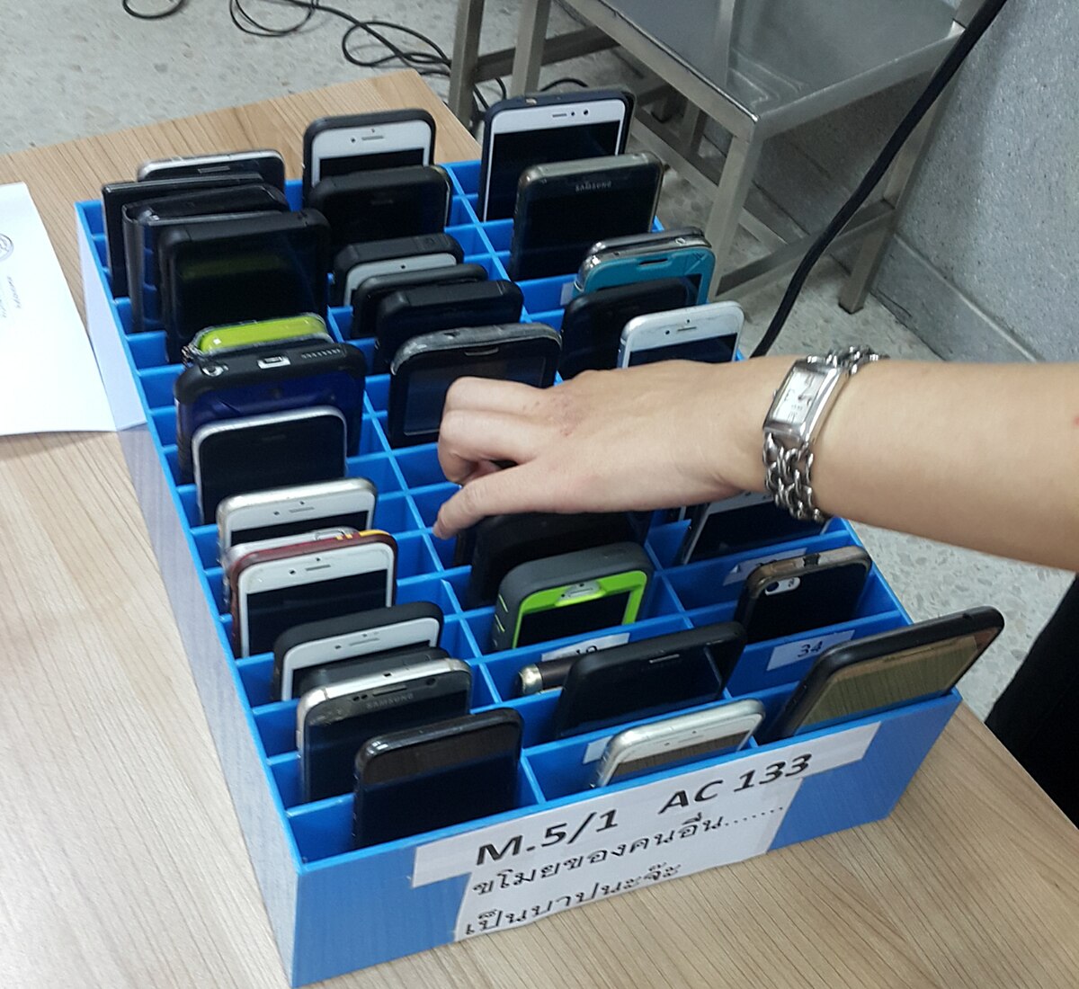 A blue plastic mobile phone cage filled with numerous smartphones, with a hand reaching in to retrieve one. The cage has labels in Thai, including "M.5/1 AC 133" and "โทรศัพท์ของคนอื่น... เป็นบาปนะจ๊ะ" (Other people's phones... it's a sin).