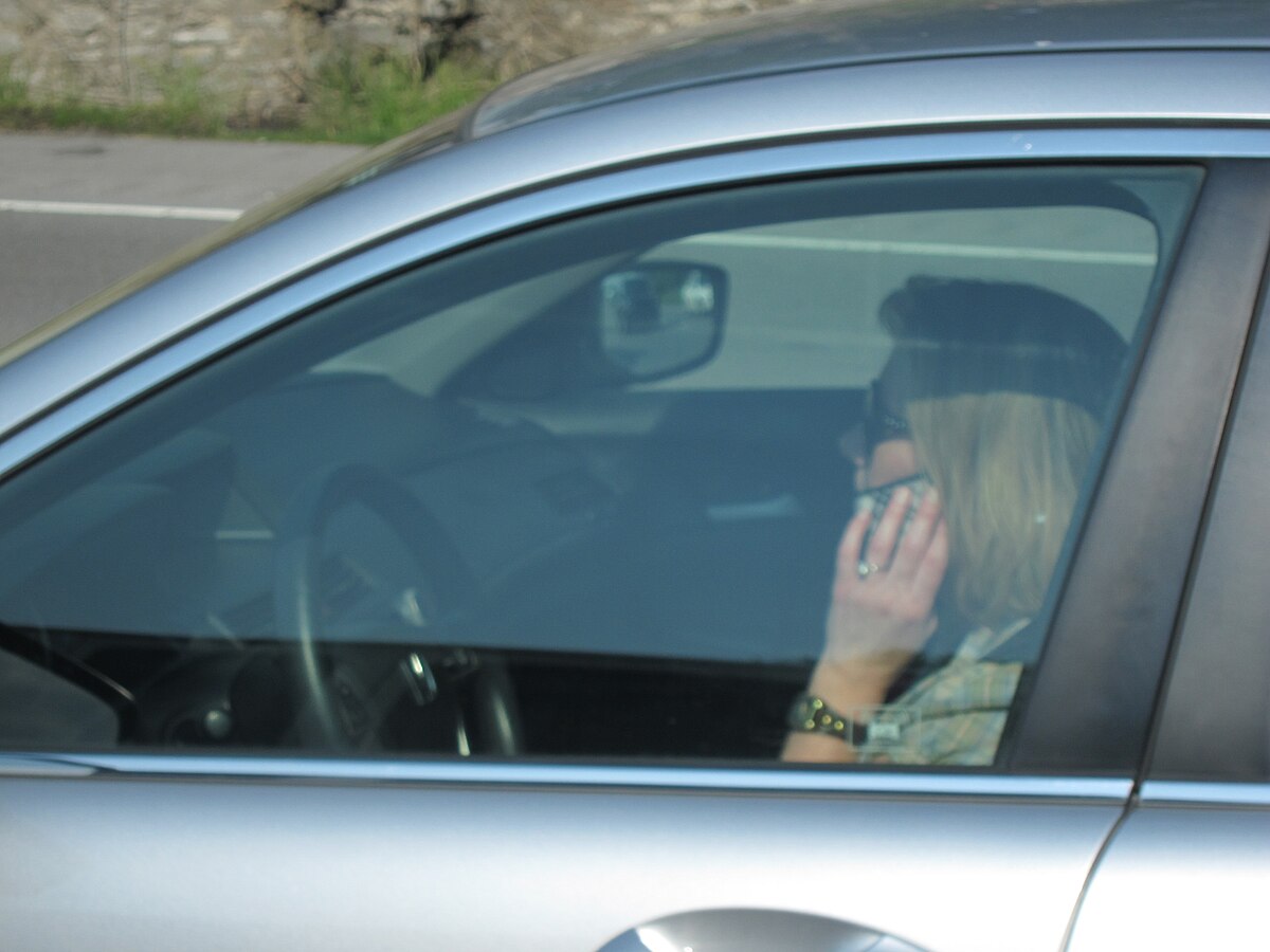 A driver, seen through the car window, is talking on a cellphone while driving. The driver's hand is holding the phone to their ear.