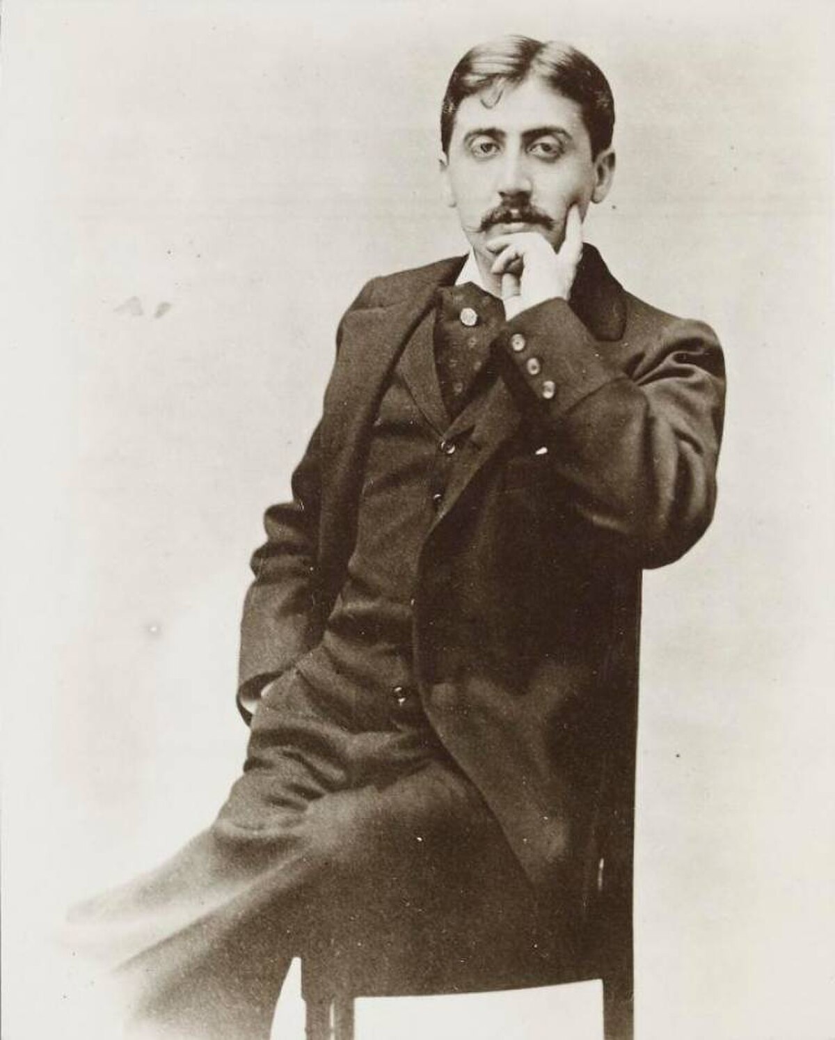 Sepia-toned portrait of Marcel Proust, taken around 1895 by Otto Wegener, showing him seated with his right hand resting on his chin. He is wearing a dark suit with a waistcoat and a tie.