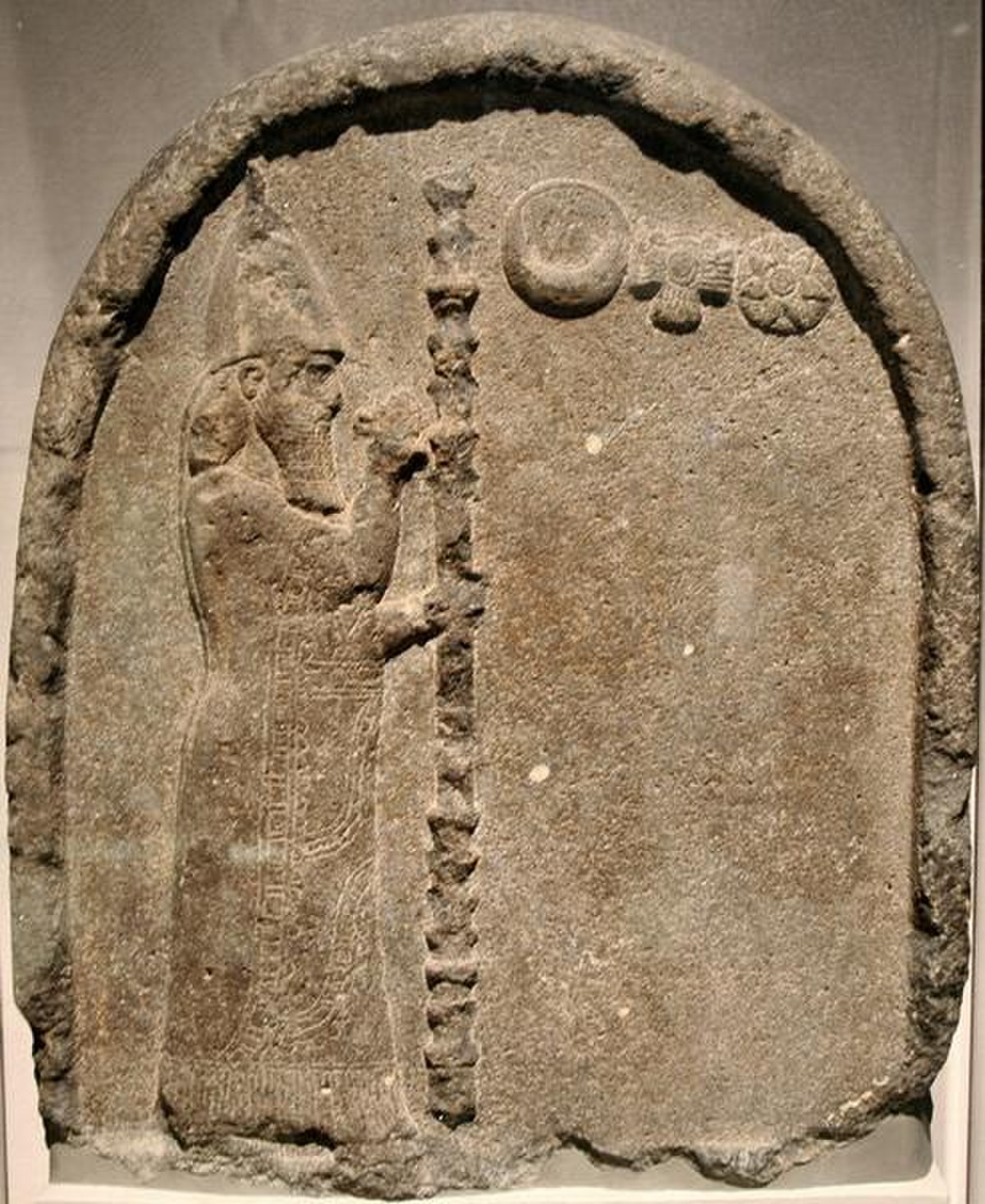A stele depicting Nabonidus, the Babylonian king, praying to celestial bodies: the moon, the sun, and the planet Venus. Nabonidus is shown in profile, wearing a conical hat and holding a staff, with cuneiform text inscribed on the right side.
