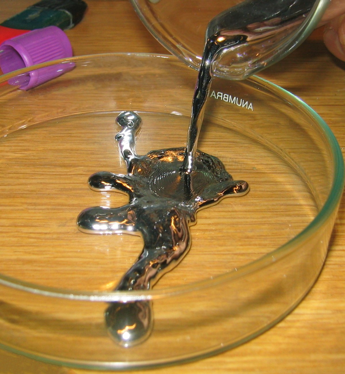 Liquid mercury is being poured from a glass beaker into a shallow glass petri dish, forming a collection of silvery, reflective droplets and puddles. The mercury exhibits its characteristic metallic sheen and high surface tension.