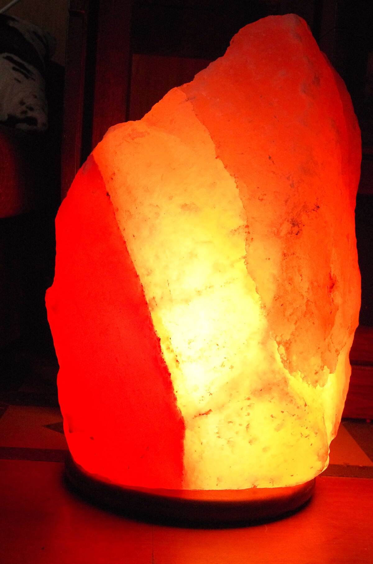 A glowing Himalayan salt lamp, carved from a large piece of pinkish-orange salt, emitting a warm, reddish-orange light. The lamp sits on a dark wooden base.