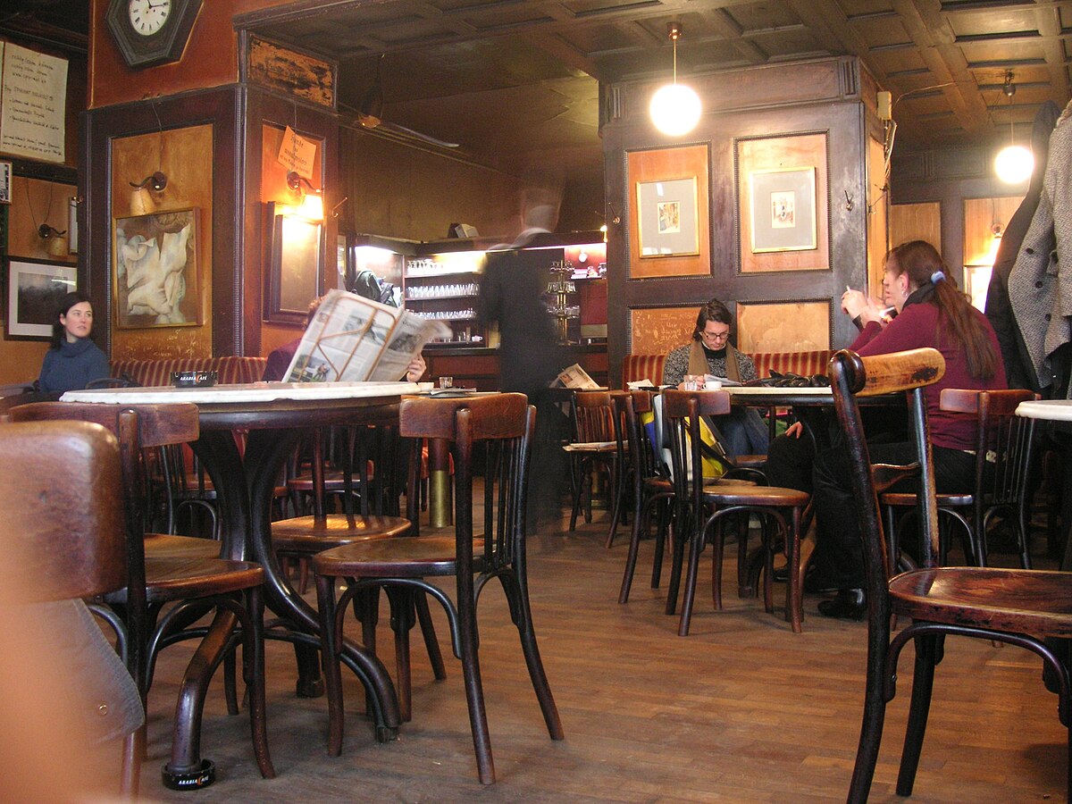 The interior of Café Hawelka in Vienna on a quiet morning, showing wooden tables and chairs, patrons reading newspapers, and a waiter in the background.