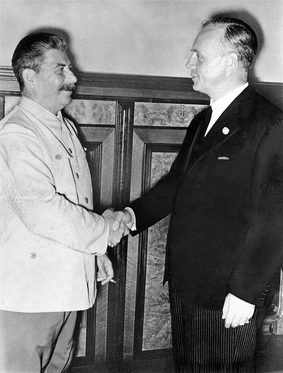 German Foreign Minister Joachim von Ribbentrop (right) and Soviet leader Joseph Stalin are shown shaking hands after signing the Molotov–Ribbentrop Pact on August 23, 1939. The two men are standing in a formal room, with Stalin wearing a light-colored uniform and Ribbentrop in a dark suit.