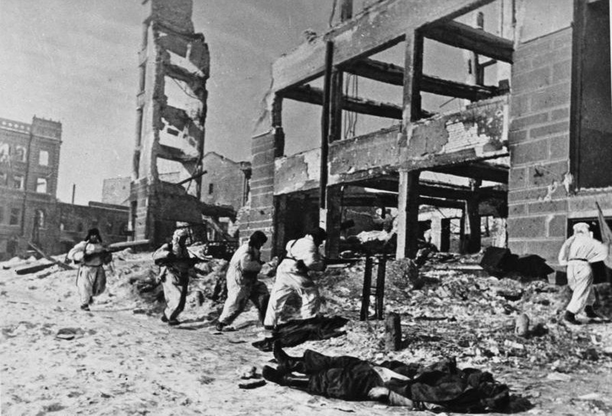 Soviet soldiers in winter camouflage move through the ruins of Stalingrad during the Kesselschlacht Stalingrad (Battle of Stalingrad). Bodies lie on the ground amidst the rubble of bombed-out buildings, indicating the harsh conditions of the battle.