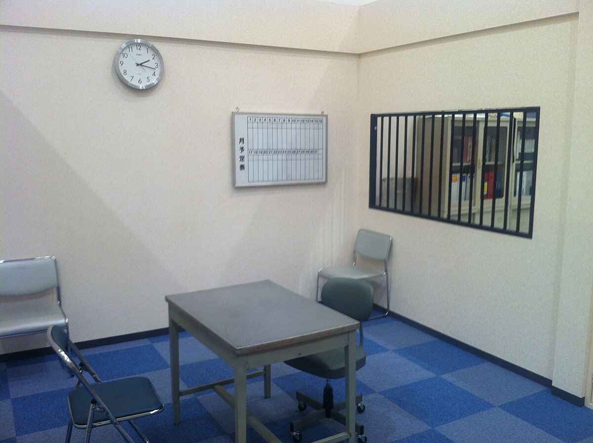 A film set designed to resemble a Japanese police interrogation room, featuring a plain room with a table, two chairs, and a clock on the wall. A barred window looks into an adjacent room, and a white board with Japanese writing is mounted on the wall.