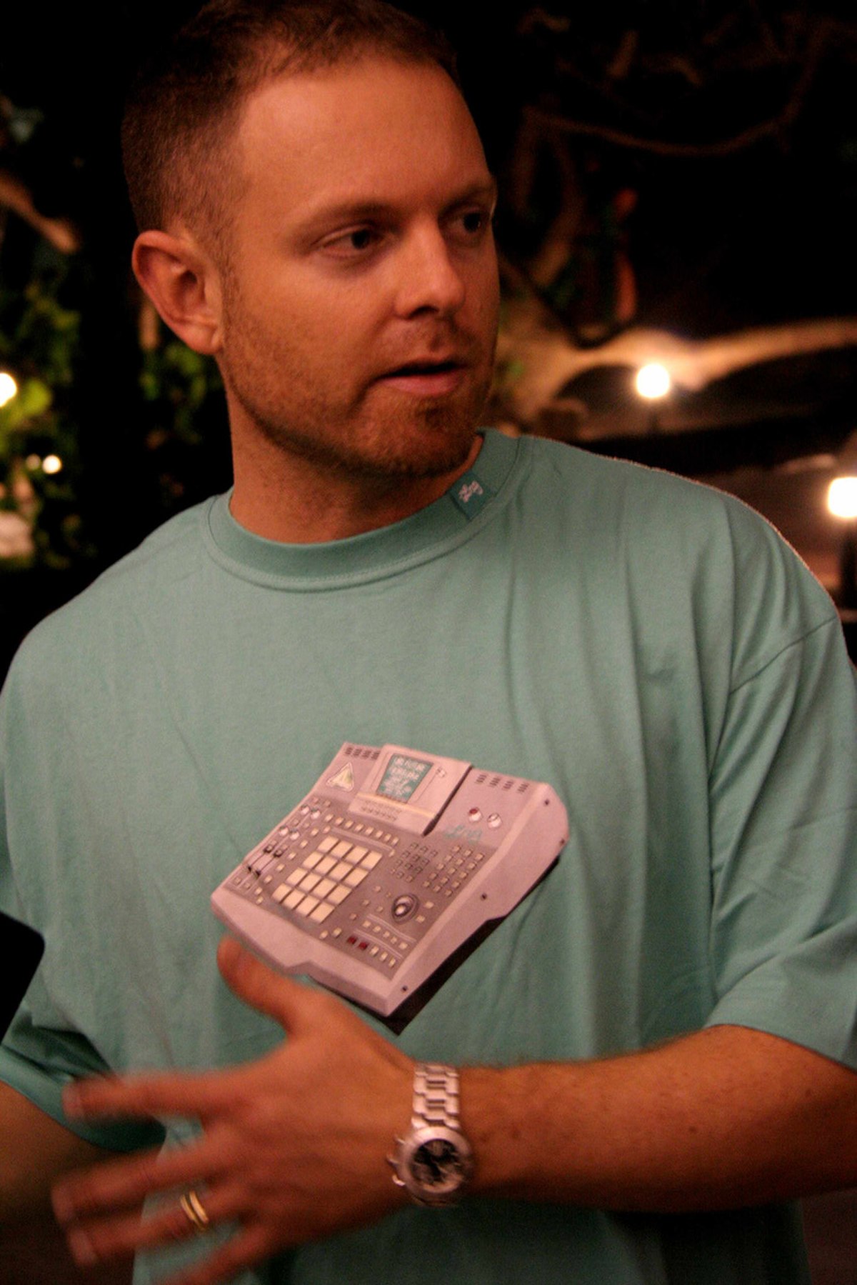 DJ Shadow, known for his 1996 album "Endtroducing" which was created entirely from samples, is shown in a close-up portrait. He is wearing a light blue t-shirt with an image of a sampler on it.