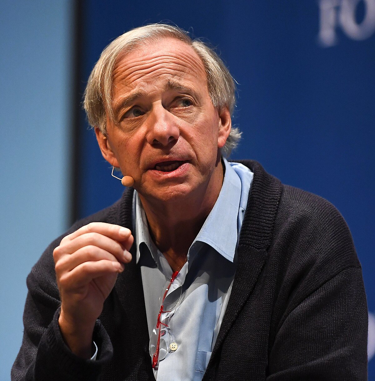 Portrait of Ray Dalio, fund manager of Bridgewater Associates, speaking at the Web Summit 2018. He is wearing a light blue shirt and a dark blue cardigan, with a microphone attached to his collar.