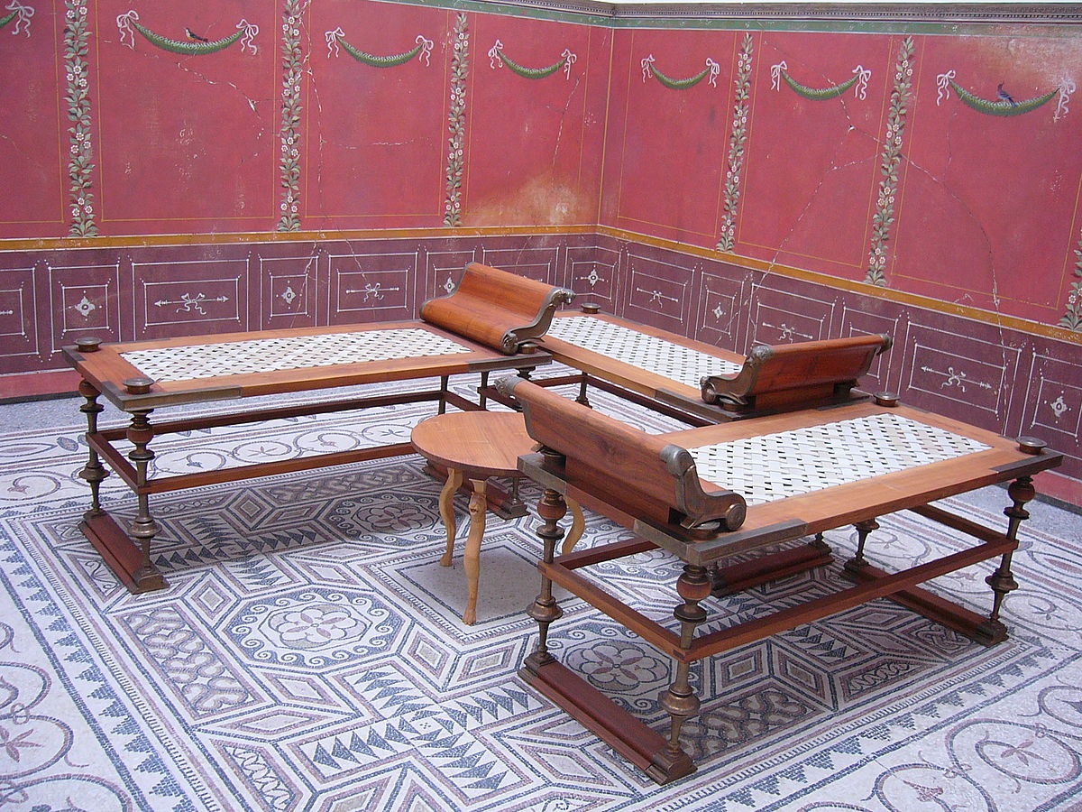 A reconstructed Roman triclinium or dining room, featuring three klinai (couches) arranged around a small table on a geometric mosaic floor. The walls are decorated with red frescoes and painted garlands.