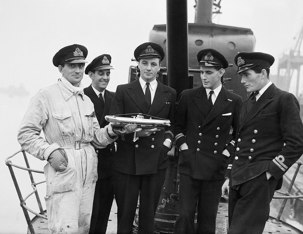 A black and white photograph of five officers from HMS Seraph standing on the deck of the submarine in December 1943. One officer in the center is holding a small, torpedo-shaped object.
