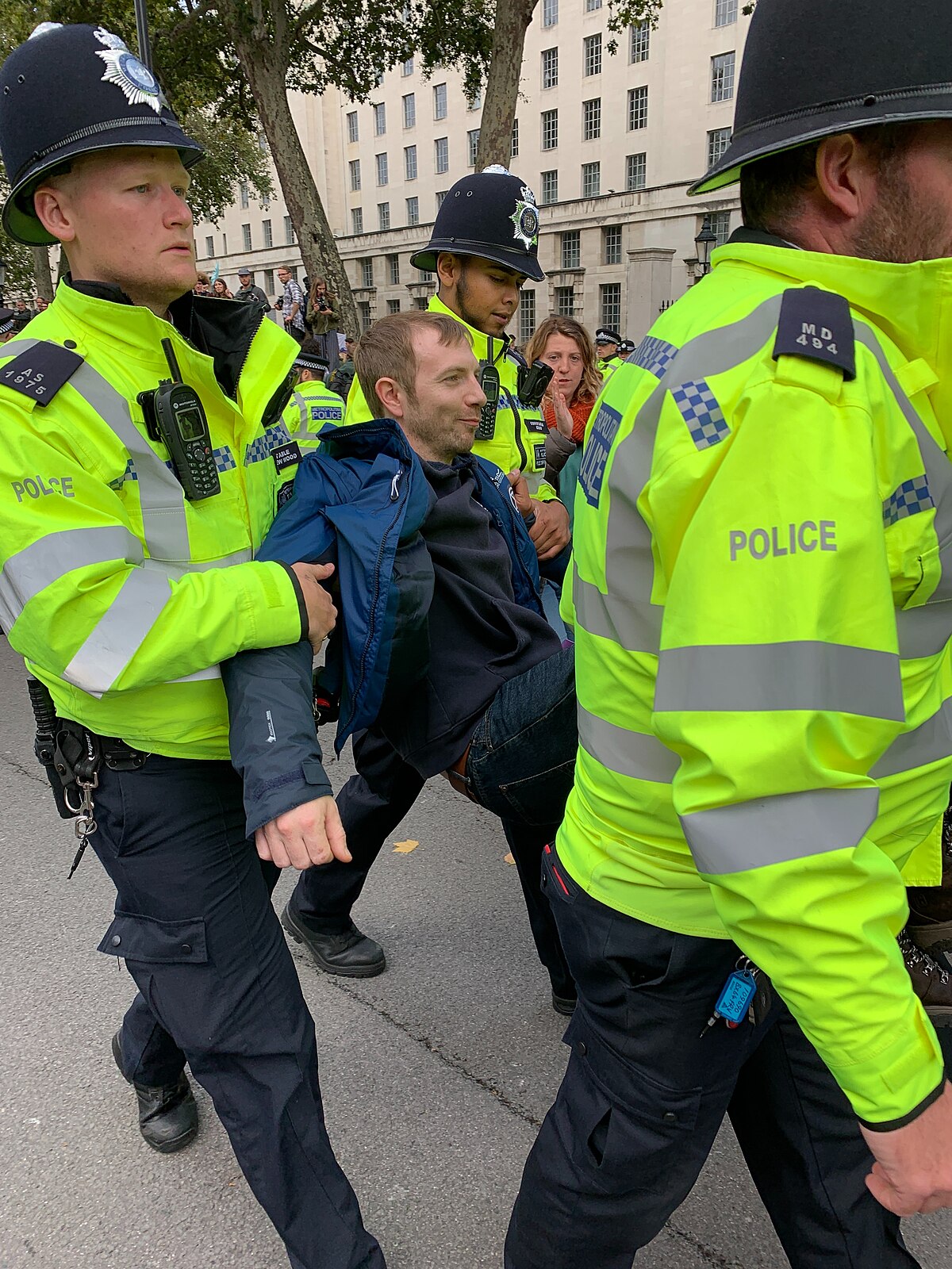 Two police officers in high-visibility vests are arresting a male Extinction Rebellion protester in Whitehall, London, during October 2019. The protester is being held by his arms and legs, while other officers and bystanders are visible in the background.