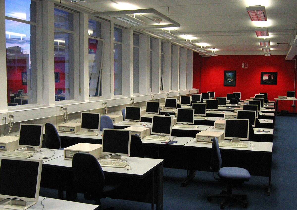 A university computer lab containing many desktop PCs arranged in rows. The room features large windows along one wall and red walls on the opposite side.
