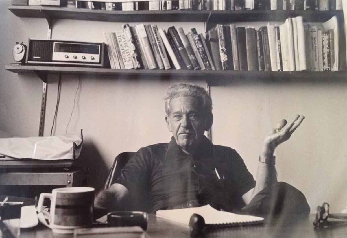 Portrait of Bertram Forer, seated at a desk with books on a shelf behind him, gesturing with his left hand. He is wearing a dark short-sleeved shirt and has a mug and other items on his desk.