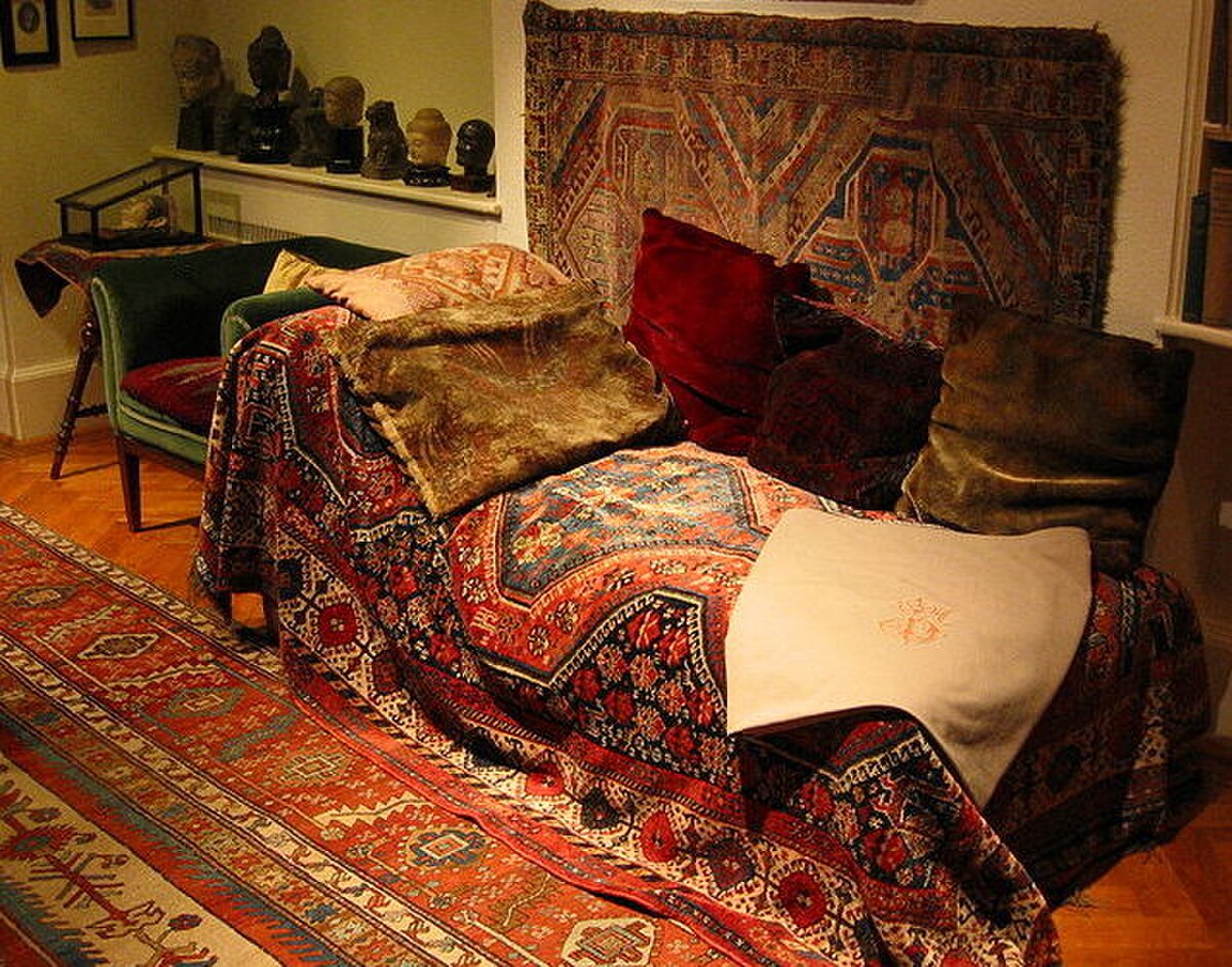 Sigmund Freud's psychoanalytic couch, covered with a richly patterned oriental rug and several cushions, located in a room with other artifacts. The couch is positioned against a wall, with a green velvet chaise lounge visible to its left.