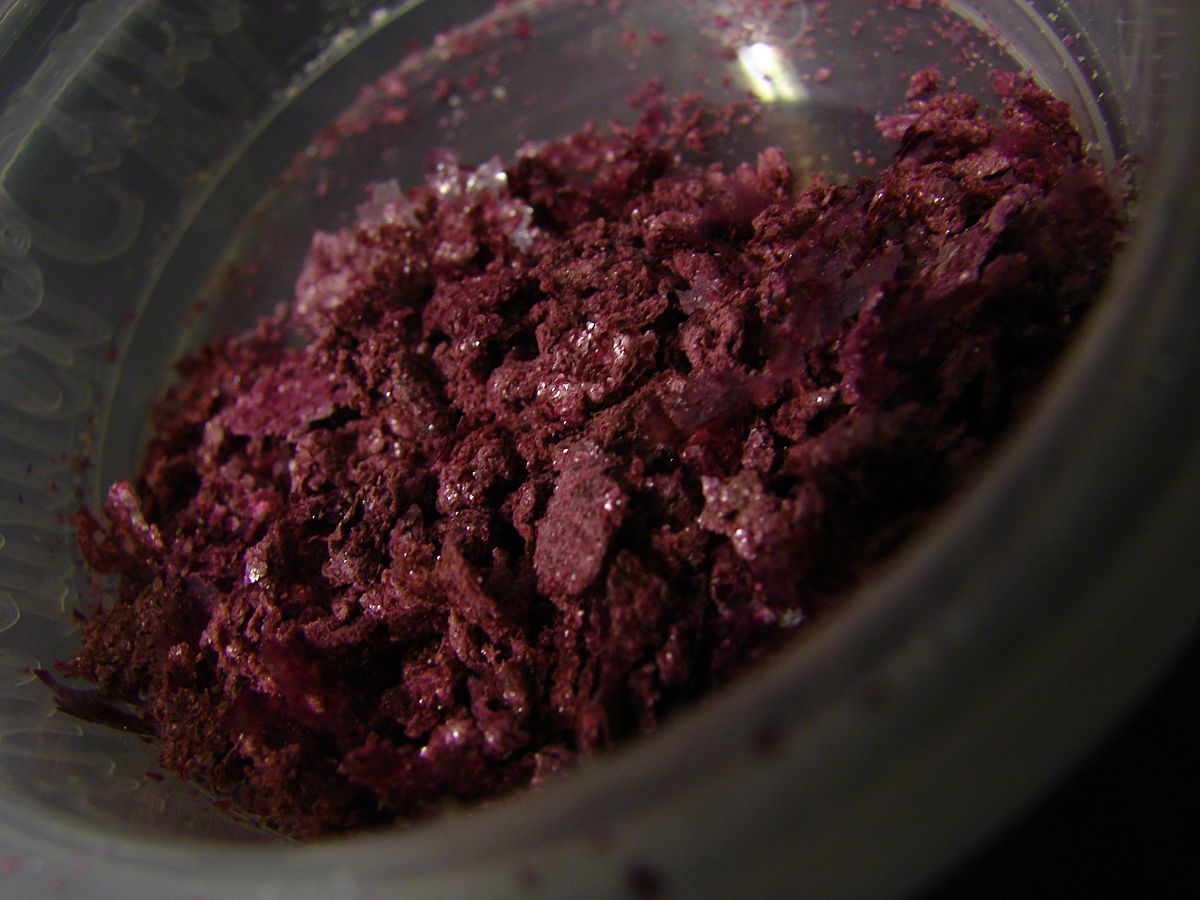 A close-up of tartaric crystals, dyed a deep reddish-purple color, in a clear plastic container. The crystals appear flaky and granular.