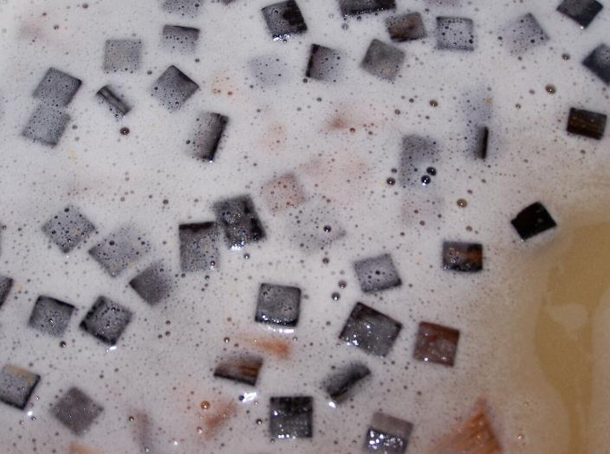 Close-up of oak chips floating in a foamy liquid, likely Chardonnay undergoing malolactic fermentation. The square-shaped oak chips are dark brown, surrounded by white foam with small bubbles.