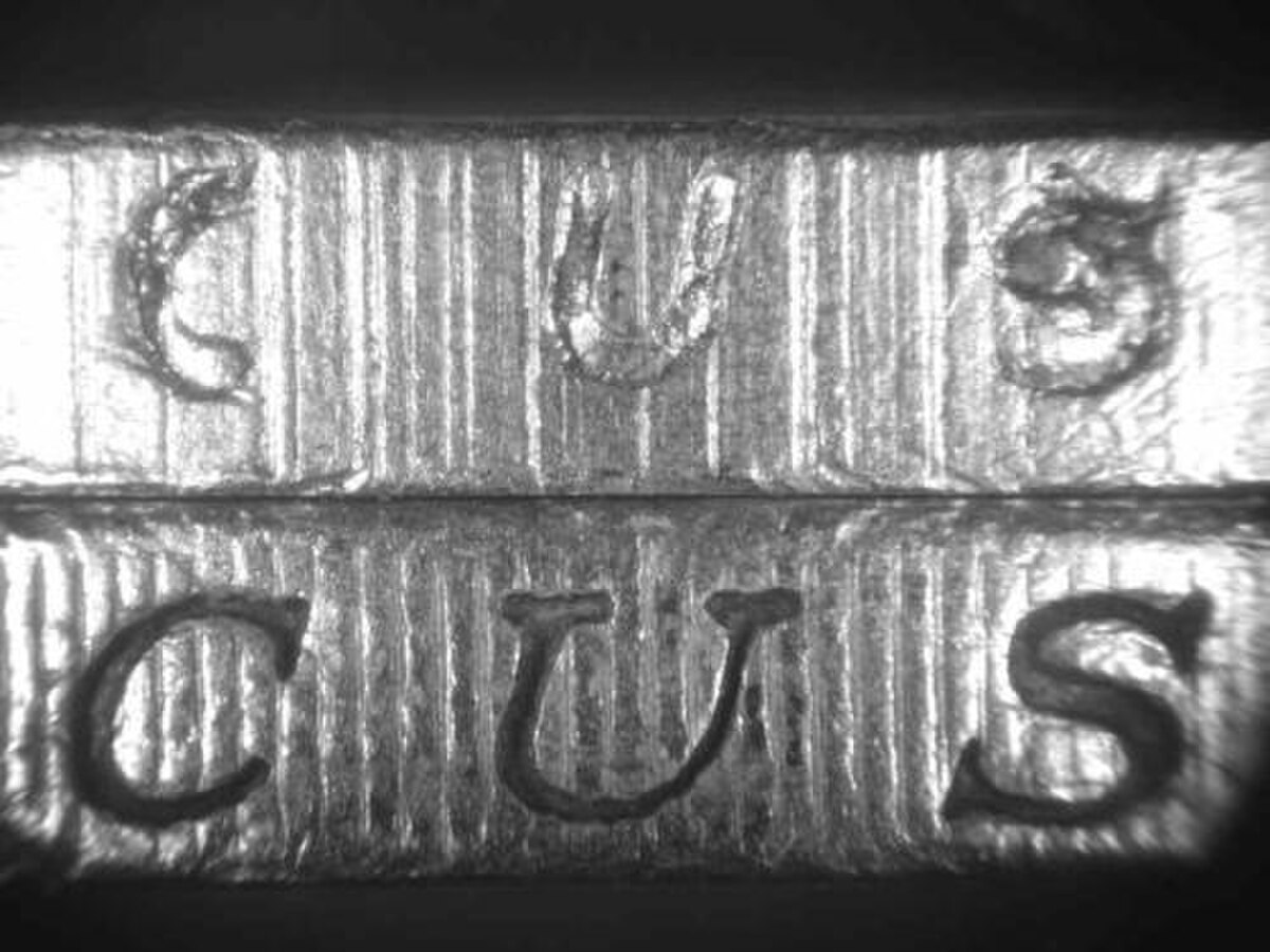 A magnified view of the edge of two coins, illustrating defective milling and letters on a counterfeit coin (top) compared to a genuine coin (bottom). The letters "CUS" are visible on both coin edges.