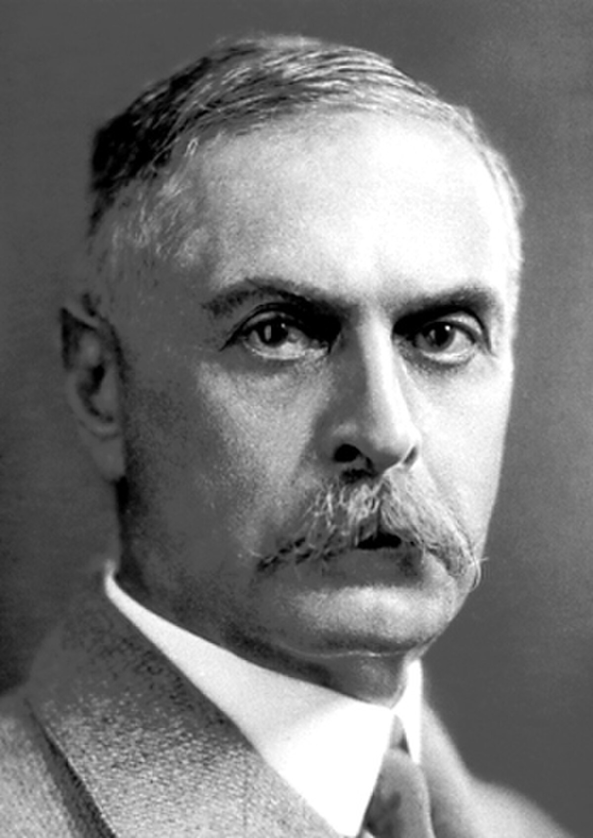 A black and white portrait of Karl Landsteiner shows him with a mustache and a serious expression, wearing a suit and tie. He is looking slightly to his right.