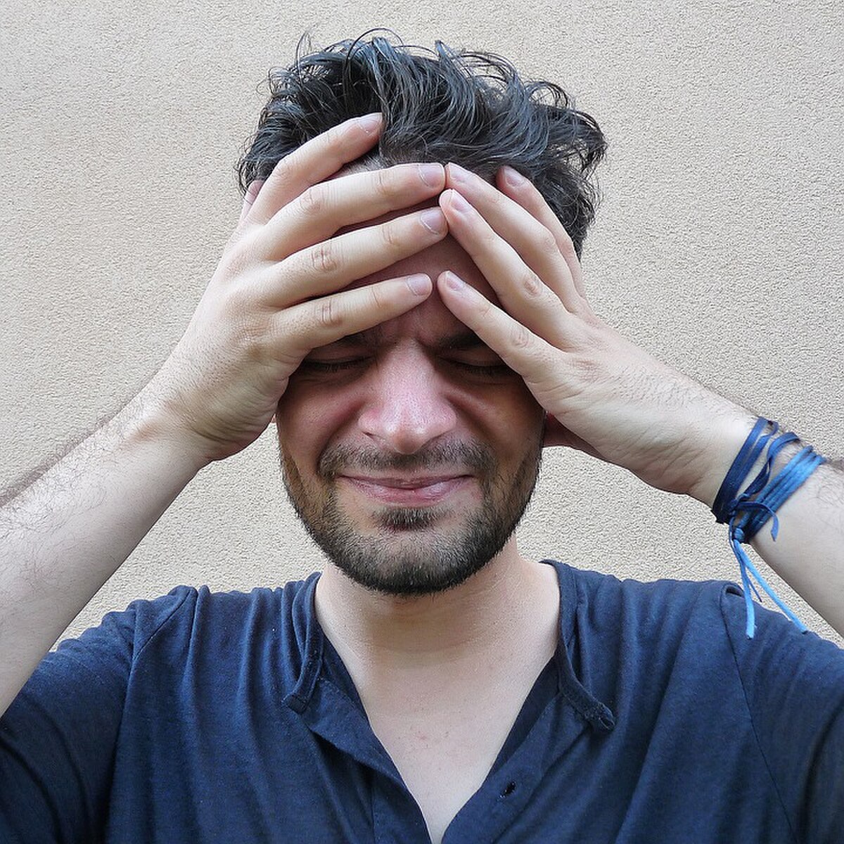 A man with dark, curly hair and a beard holds his head in his hands, with his eyes closed and a pained expression, indicating a headache or stress. He is wearing a dark blue t-shirt, and a light-colored wall serves as the background.