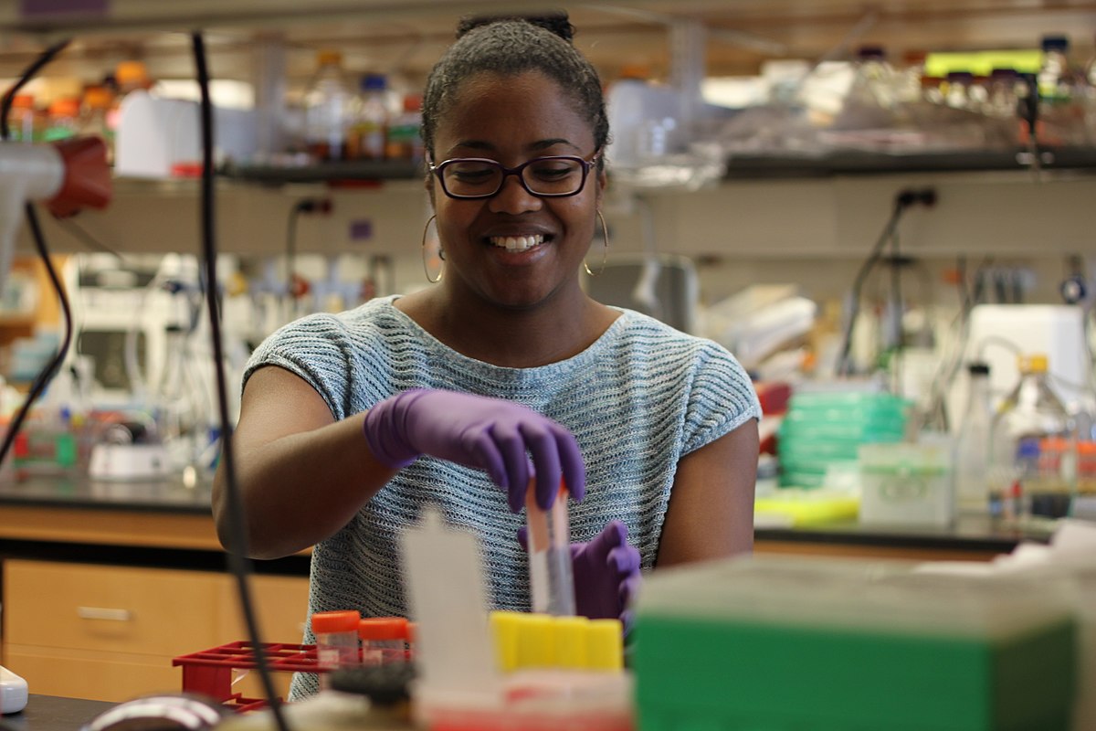 A biochemist wearing glasses and purple gloves smiles while working with test tubes and laboratory equipment. She is engaged in bench research in a brightly lit laboratory setting.