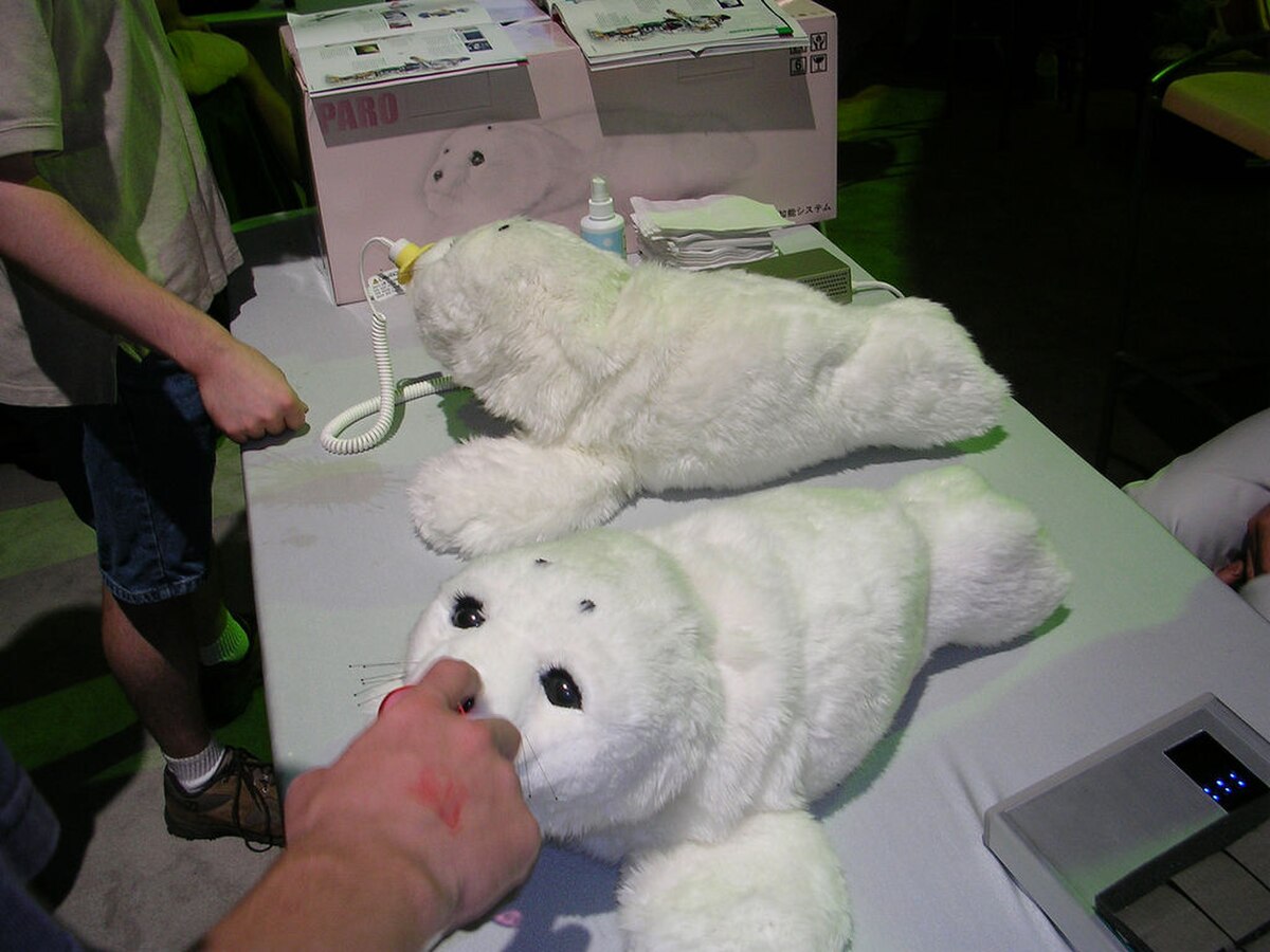 Two white robotic seal pets, named Paro, are displayed on a table, with a person's hand interacting with one of them. Paro is classified as a medical device by U.S. regulators, designed to provide therapeutic benefits.