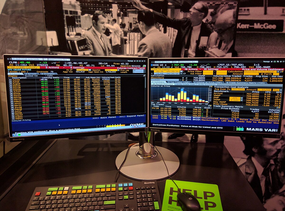 A Bloomberg Terminal setup is displayed with two monitors showing financial data and charts, and a specialized keyboard with color-coded keys. The background features a black and white image of people in a trading room, with "Ken-McGee" visible on the right.