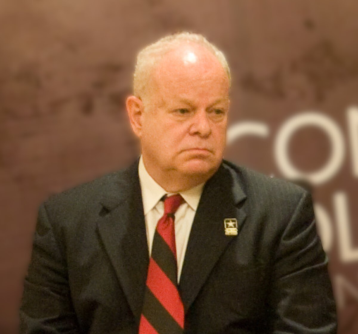 Portrait of Martin Seligman, a man with white hair and a serious expression, wearing a dark suit and a red tie. He has a small pin on his lapel.