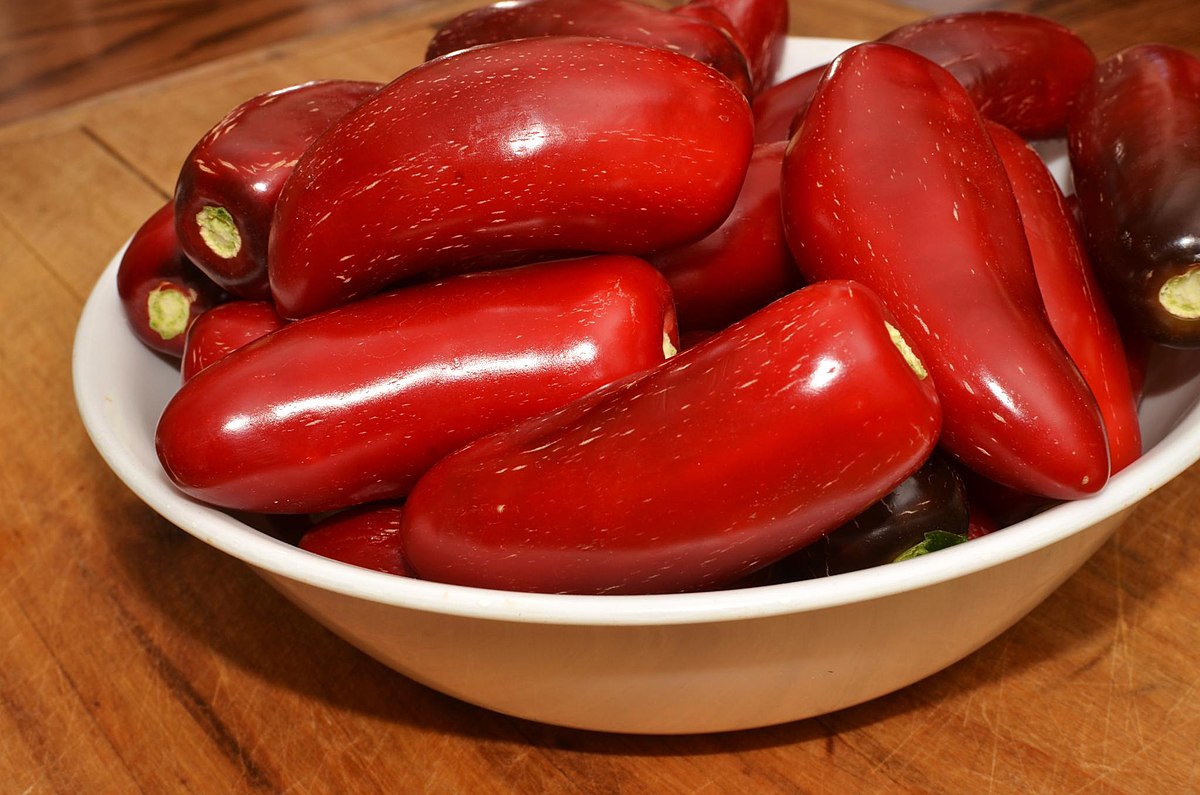 A white bowl filled with numerous ripe red jalapeño peppers, showcasing their deep red color and smooth, glossy skin. Some peppers show slight variations in color, indicating different stages of ripeness.