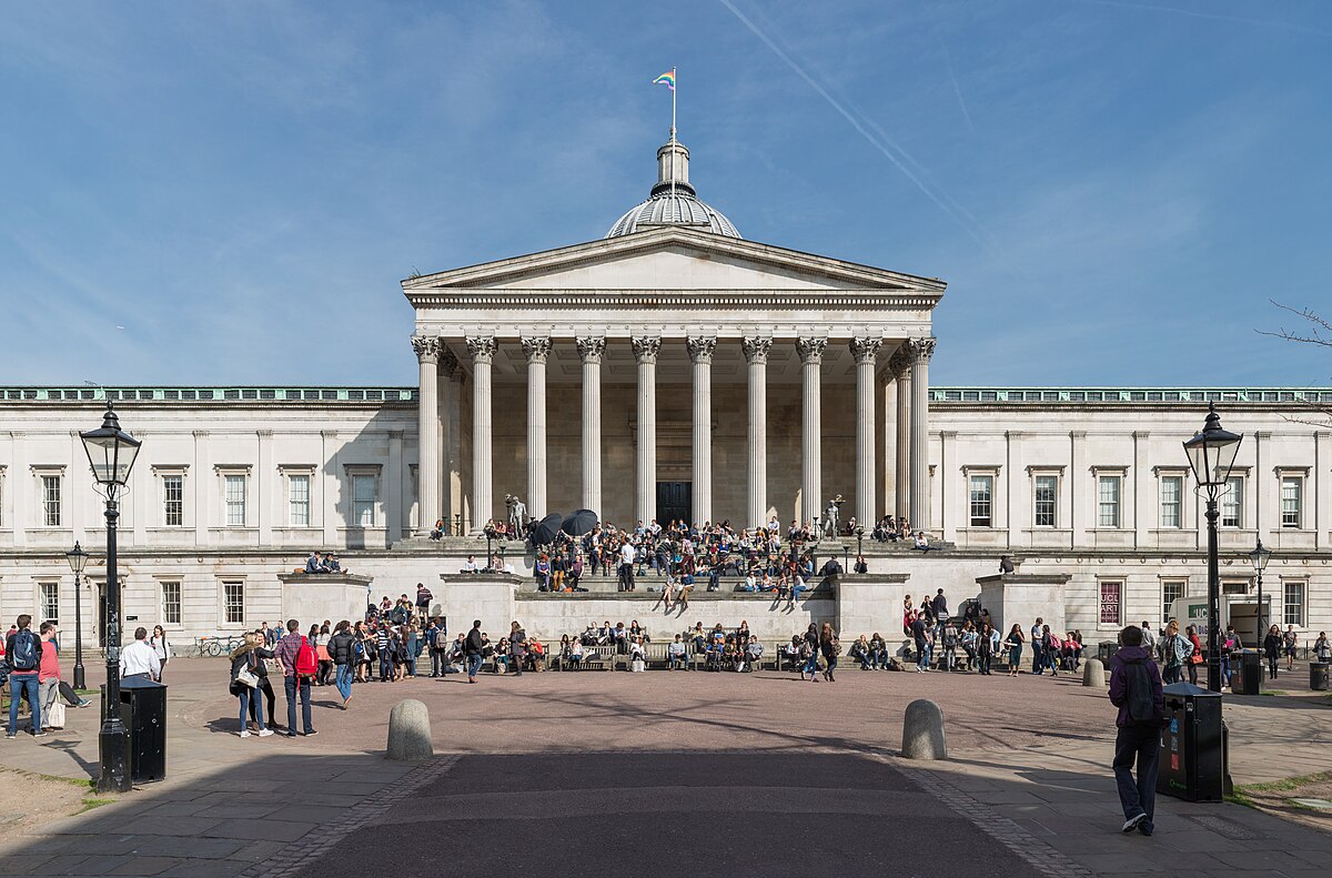 The Wilkins Building at University College London (UCL), featuring its iconic neoclassical portico and dome, with numerous students and visitors gathered on the steps and surrounding plaza on a sunny day. The building is a prominent feature of the UCL campus.