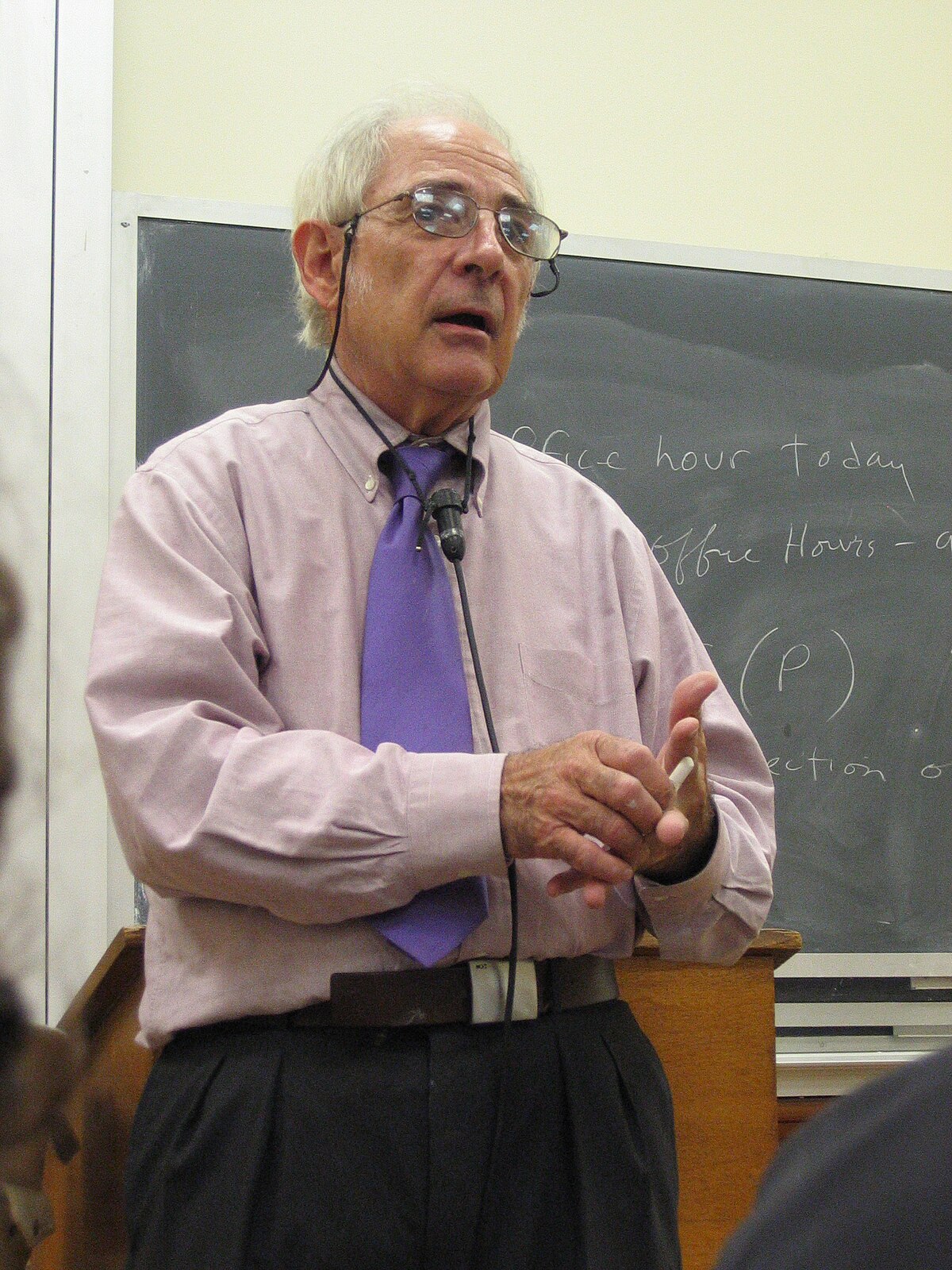 Portrait of philosopher John Searle speaking in December 2005, wearing a light purple shirt, a dark purple tie, and glasses. He is gesturing with his hands in front of a blackboard with some writing on it.
