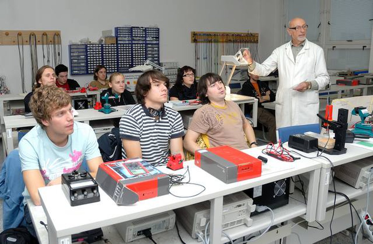 A lecture is taking place at the Faculty of Biomedical Engineering, CTU in Prague, with a male instructor in a white lab coat addressing a classroom of students. The students are seated at desks equipped with various electronic devices and equipment.
