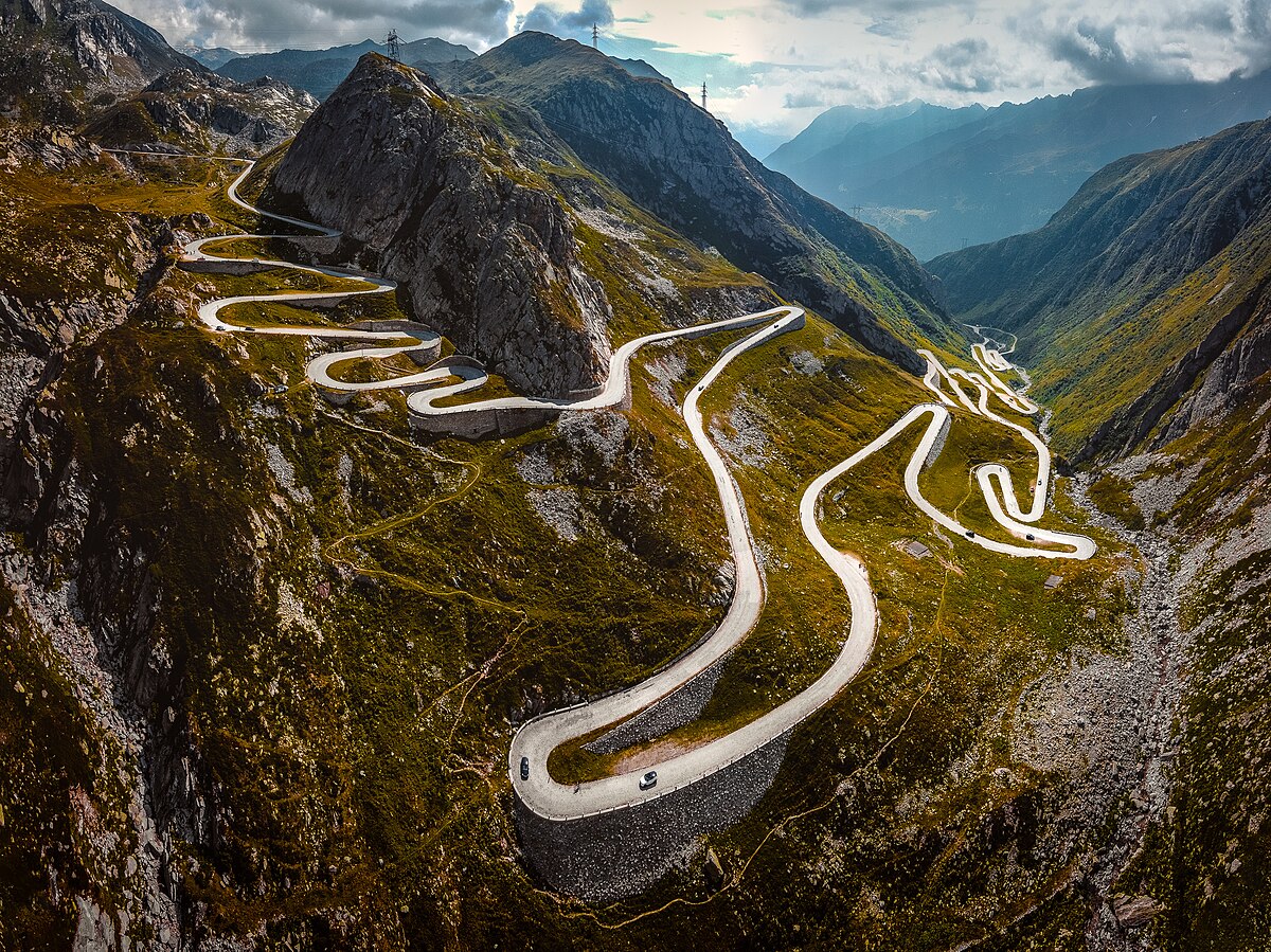 An aerial view of the Old Tremola Road, a winding serpentine road traversing a mountainous landscape in Switzerland. The road features numerous hairpin turns as it ascends and descends the steep terrain, with green vegetation and rocky outcrops visible on either side.