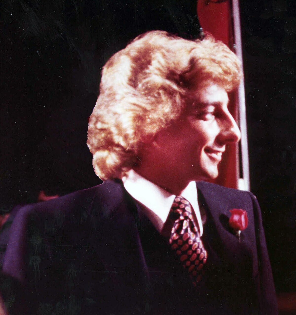 A portrait of Barry Manilow from 1979, showing him in profile, smiling, wearing a dark suit with a patterned tie and a red rose boutonnière. He has curly blonde hair and is looking to the right.