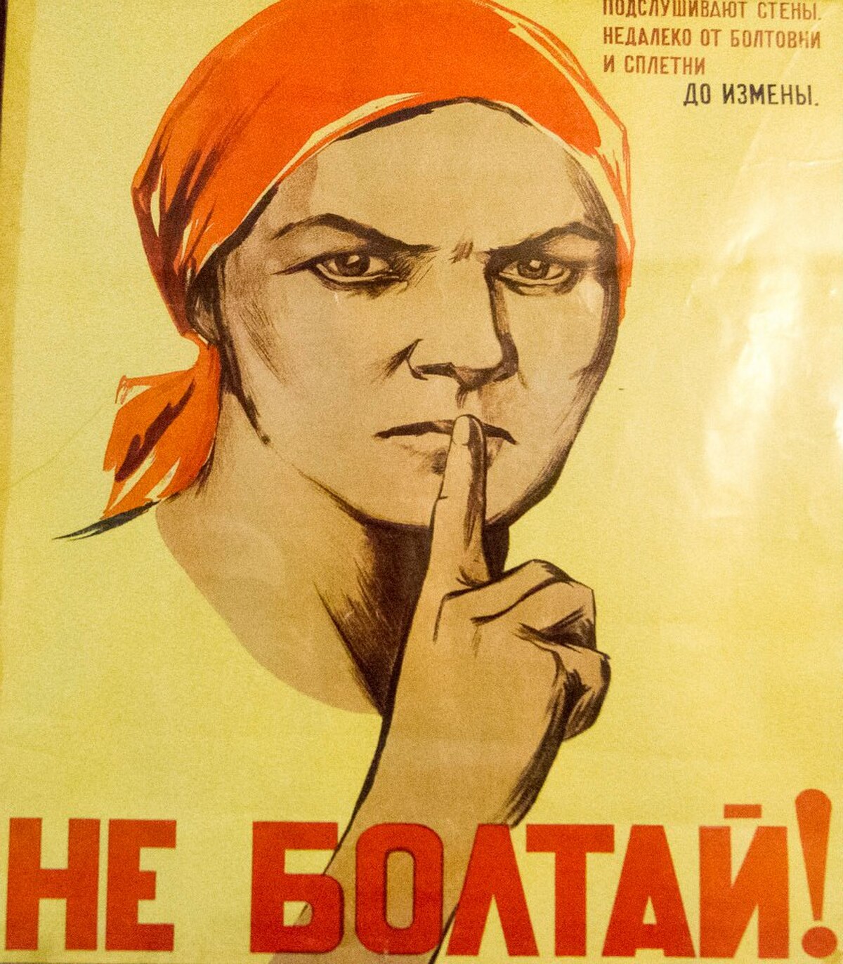 A Soviet propaganda poster featuring a stern-faced woman in a red headscarf holding a finger to her lips in a gesture of silence. The Russian text at the bottom reads "НЕ БОЛТАЙ!" (Ne Boltai!), meaning "Don't Chatter!", and the smaller text above warns against loose talk leading to treason.