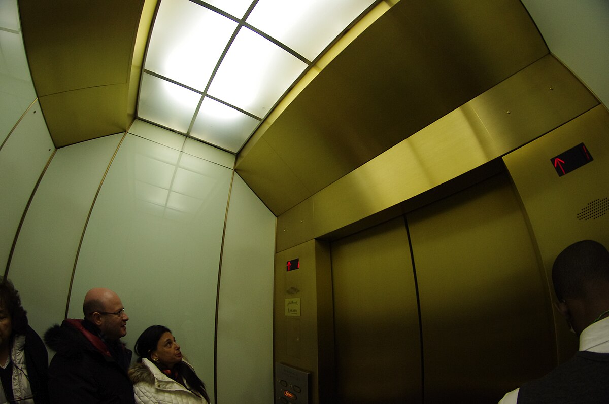 The interior of an Otis Elevator Company express elevator, featuring golden walls and ceiling, serving the upper floors of the 875 North Michigan Avenue building. Passengers are visible inside the elevator, which provides access to the 94th floor observation deck, 95th floor restaurant, and 96th floor cocktail lounge.