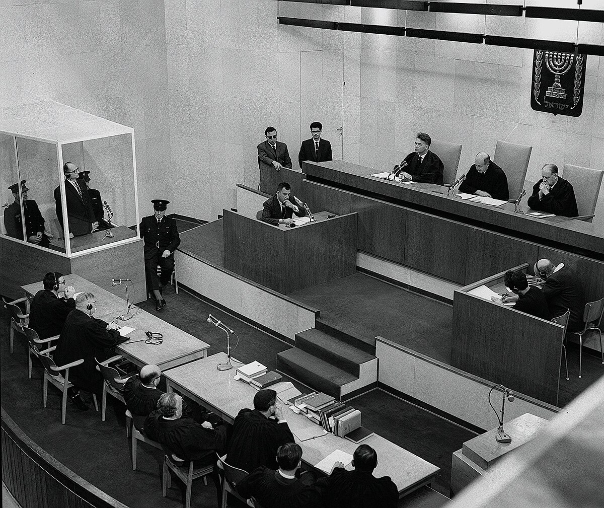 Adolf Eichmann is shown in a glass booth during his sentencing at the conclusion of the Eichmann Trial. The courtroom is filled with legal personnel, including judges on a raised dais and lawyers at tables below.