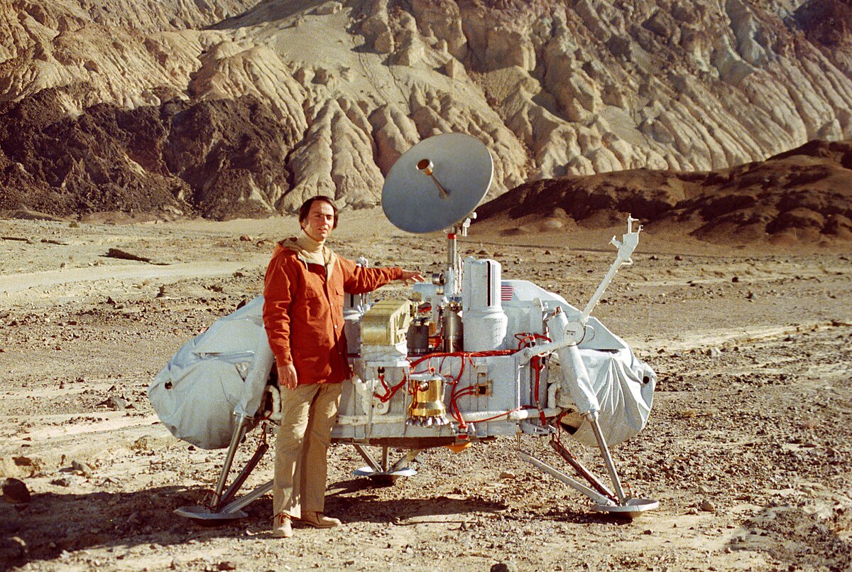 Carl Sagan stands next to a Viking lander model in a desert landscape with rocky hills in the background. He is wearing a red jacket and light-colored pants, with the lander featuring a large dish antenna.