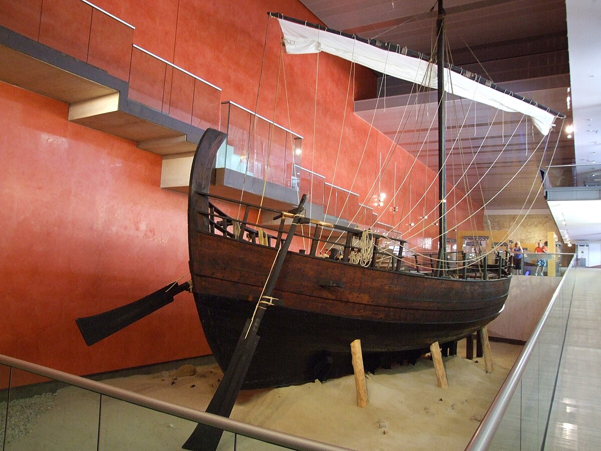 A full-scale replica of the Kyrenia ship, a 4th-century BCE Greek merchant ship, displayed indoors in a museum setting. The wooden ship features a single mast with a square sail and is surrounded by sand, with a red wall in the background.