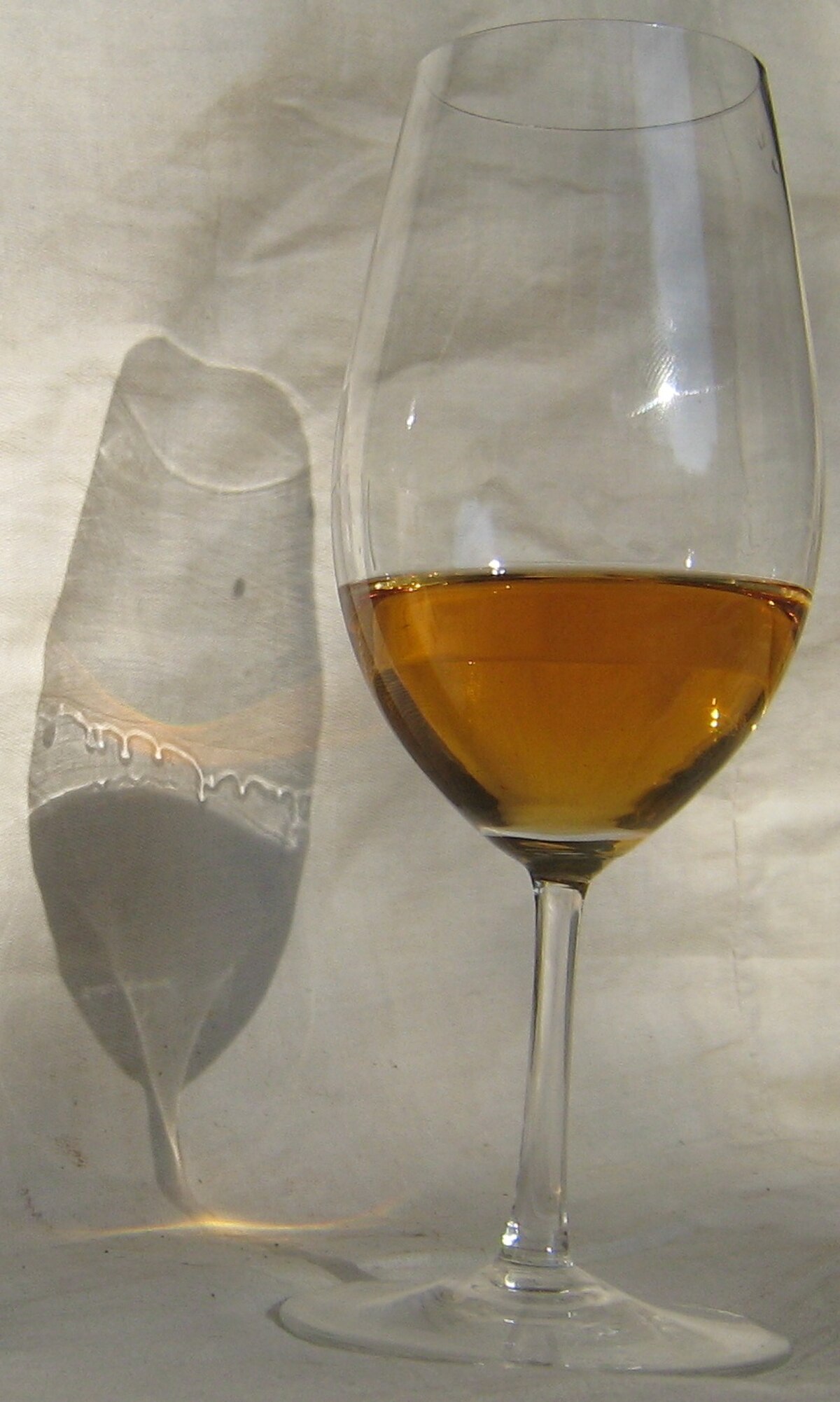 A glass of amber-colored wine with a 13.5% alcohol content is shown, casting a shadow on a textured white background. The shadow clearly displays the "tears of wine" effect, where rivulets of wine cling to the glass.